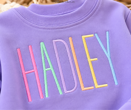 A close-up of a lavender purple sweatshirt featuring the name 'HADLEY' embroidered across the chest in pastel pink, green, purple, orange, yellow, and blue lettering.