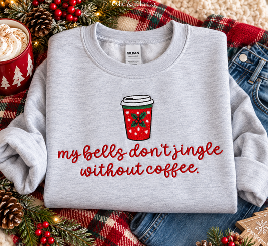 A light gray Gildan sweatshirt with a red and green holiday coffee cup graphic and the text 'my bells don't jingle without coffee.' in red script, styled in a flat lay with jeans, a plaid blanket, pinecones, and a mug of hot cocoa.