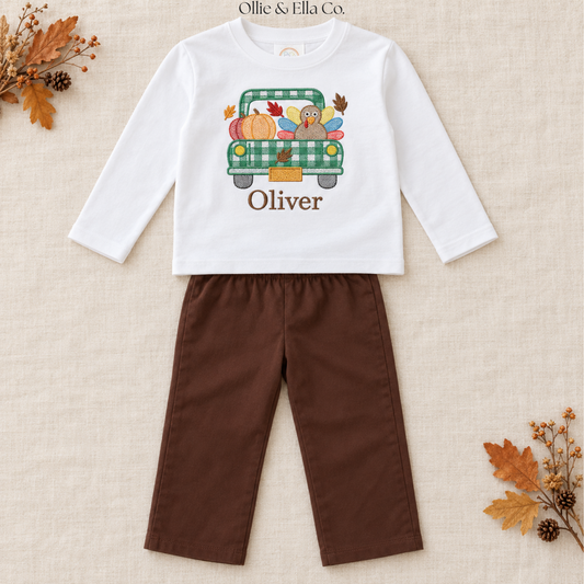 A toddler boy's two-piece outfit set by Ollie & Ella Co. featuring a white long-sleeve shirt with a green plaid truck graphic and the name 'Oliver' printed on the front, paired with brown pants, laid flat on a textured beige fabric background with autumn leaf props.