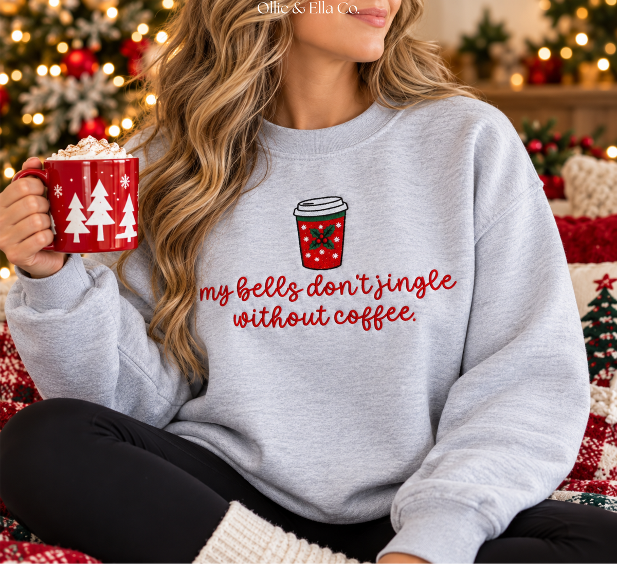 A woman wearing a light gray crewneck sweatshirt with a red graphic of a coffee cup and the text 'my bells don't jingle without coffee.' She is holding a red mug with white Christmas tree silhouettes, set against a cozy holiday background with a blurred Christmas tree and warm lights.