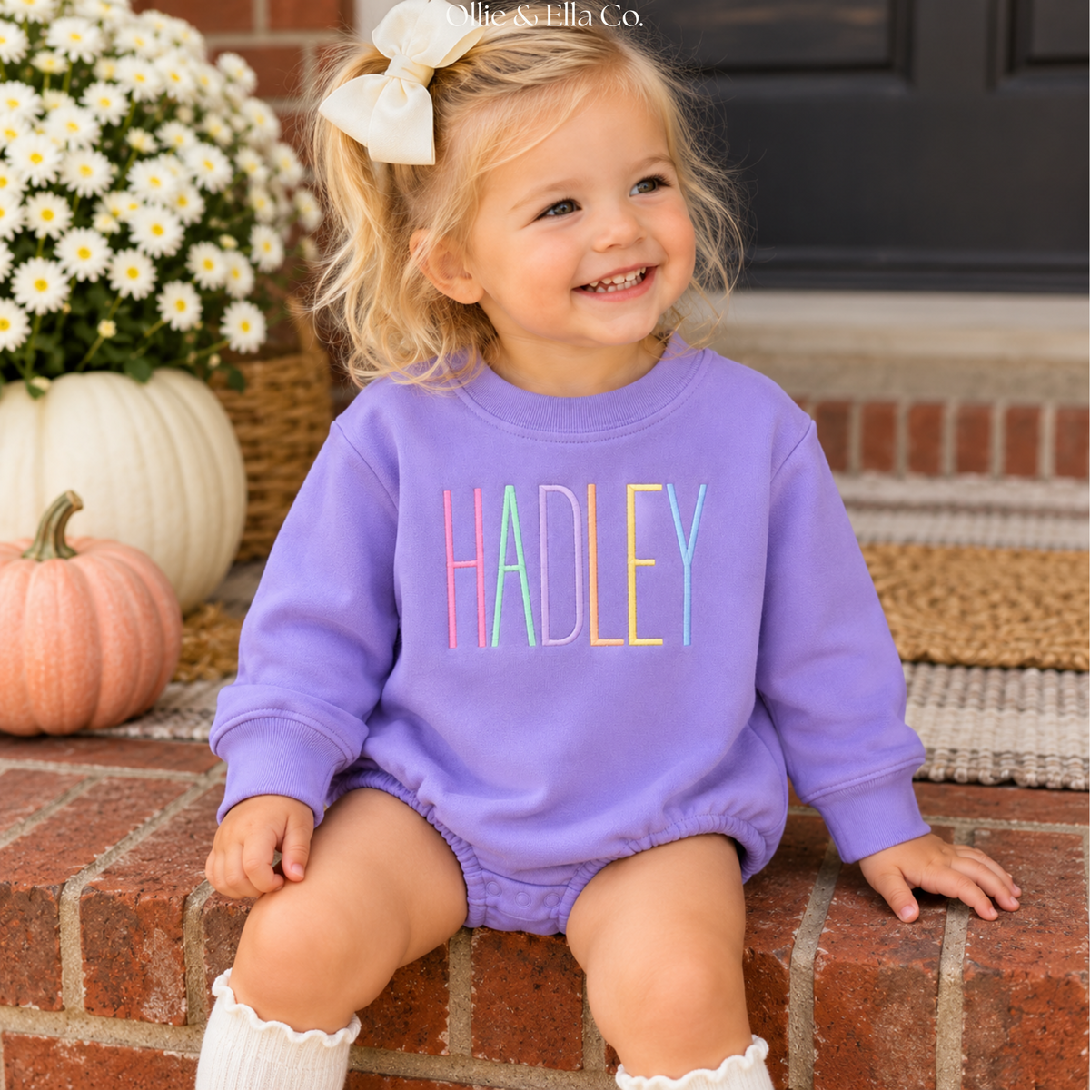 A toddler girl with a white bow sits on brick steps wearing a lavender long-sleeve romper with the name 'HADLEY' printed across the chest in colorful letters. Pumpkins and white daisies are visible in the background.