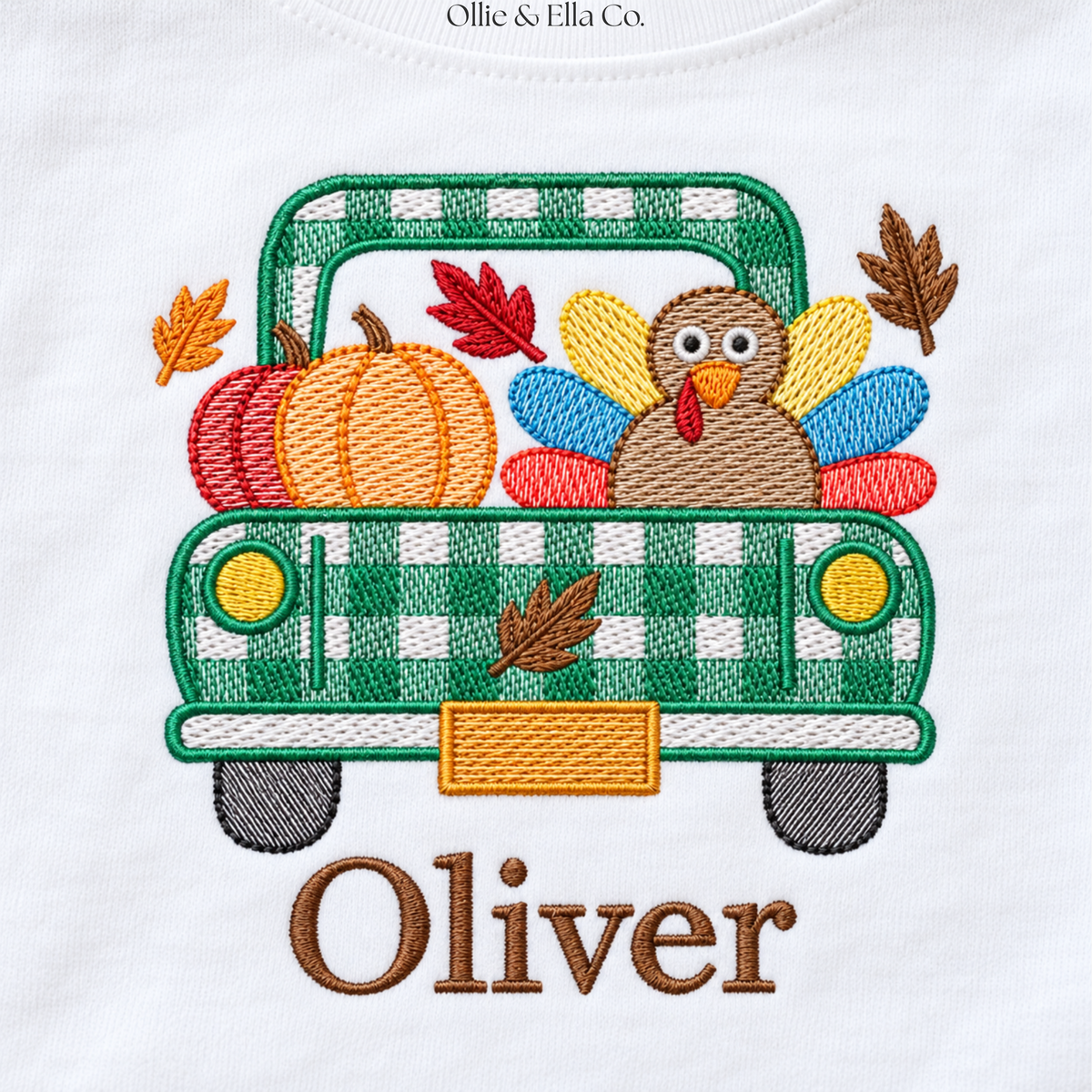 A close-up of a white garment featuring an embroidered autumn truck design with pumpkins, a turkey, leaves, and the name 'Oliver' in brown script. The top shows the text 'Ollie & Ella Co.