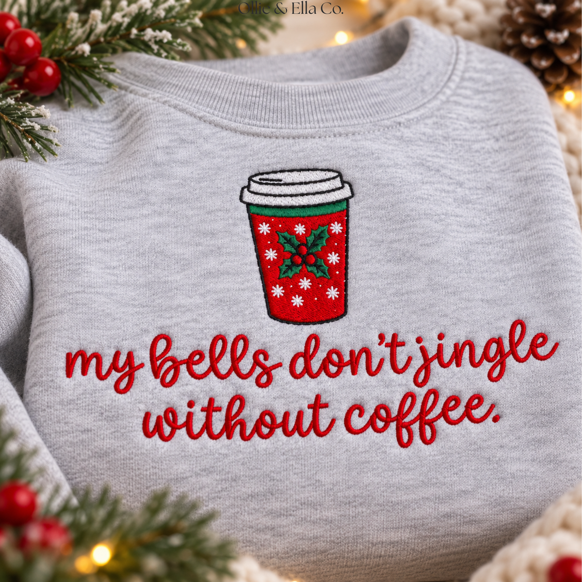 A light heather gray crewneck sweatshirt featuring a festive embroidered design of a red holiday coffee cup with green holly leaves and white text reading 'my bells don't jingle without coffee.' The sweatshirt is styled with Christmas greenery and warm string lights.