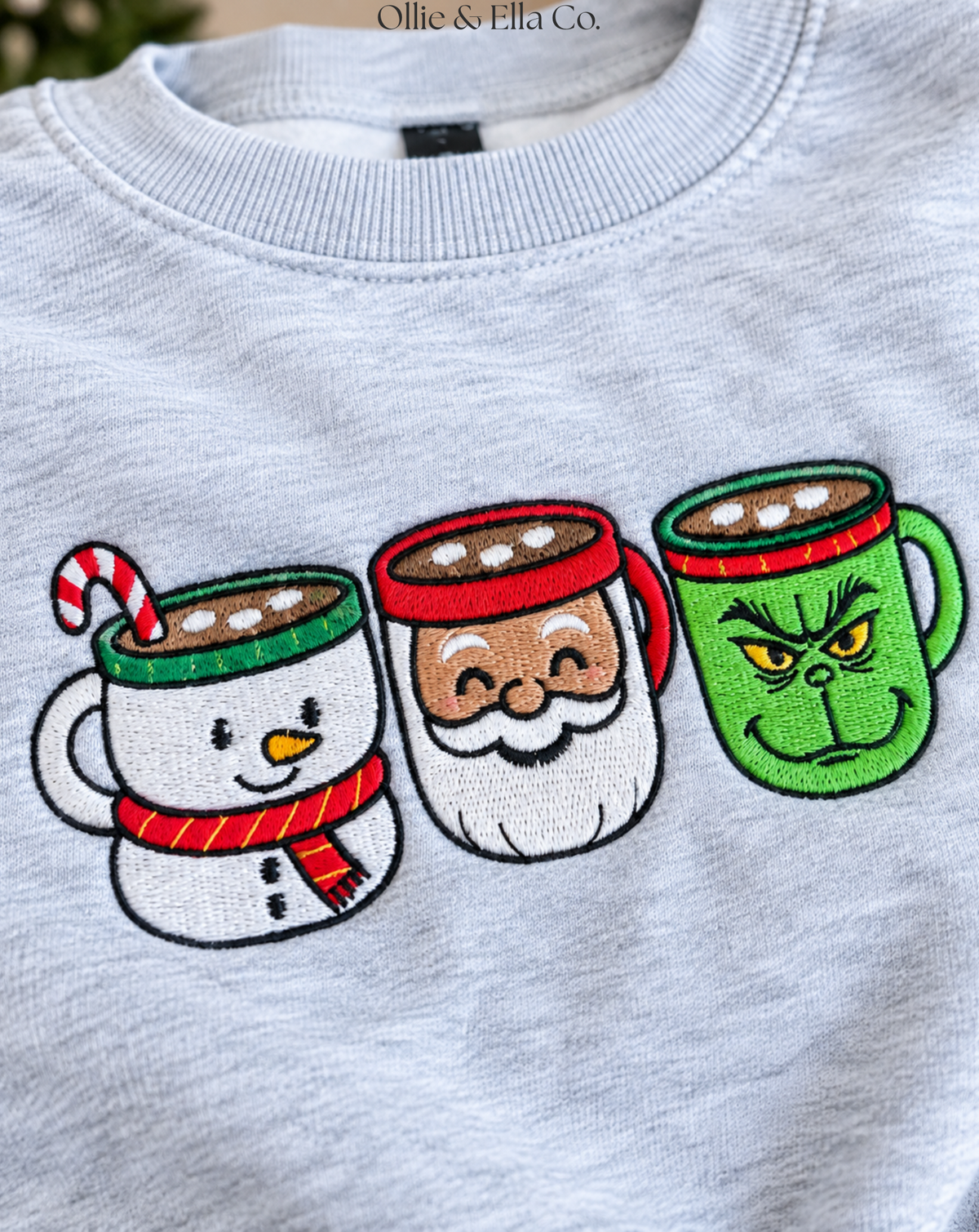 A close-up of a light gray heather crewneck sweatshirt featuring a graphic of three embroidered holiday-themed coffee mugs: a snowman with a candy cane, Santa Claus, and the Grinch.