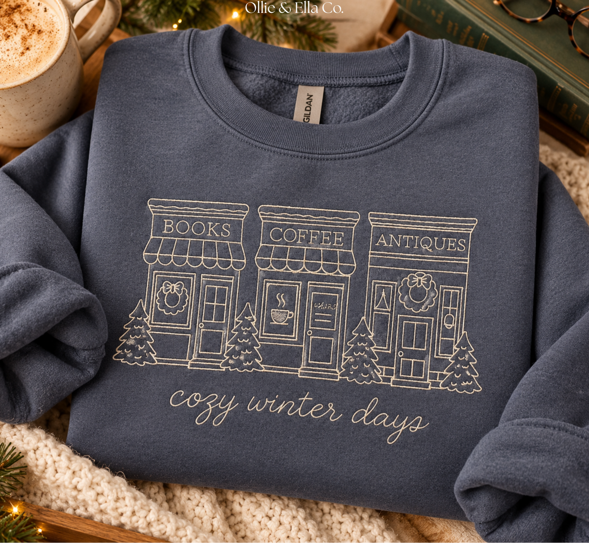 A folded navy blue sweatshirt with a white graphic of three storefronts labeled Books, Coffee, and Antiques, with the phrase 'cozy winter days' below. The sweatshirt is styled on a cozy knit blanket with a mug, book, and glasses in the background.