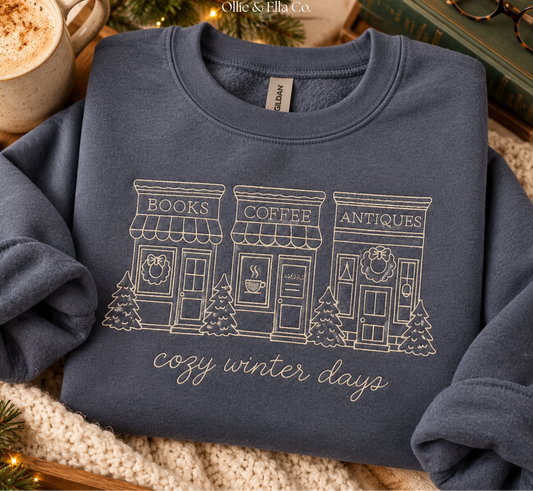 A folded navy blue sweatshirt with a white graphic of three storefronts labeled Books, Coffee, and Antiques, with the phrase 'cozy winter days' below. The sweatshirt is styled on a cozy knit blanket with a mug, book, and glasses in the background.
