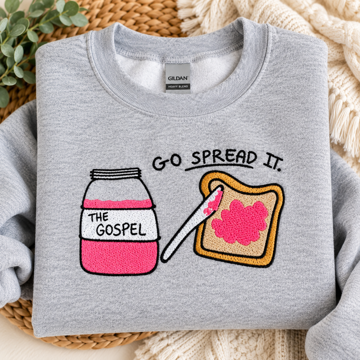 A light gray crewneck sweatshirt laid flat on a woven mat with a cream knit blanket. The sweatshirt features a graphic of a pink-and-white jar labeled 'THE GOSPEL' next to a slice of toast with pink spread and the text 'GO SPREAD IT.