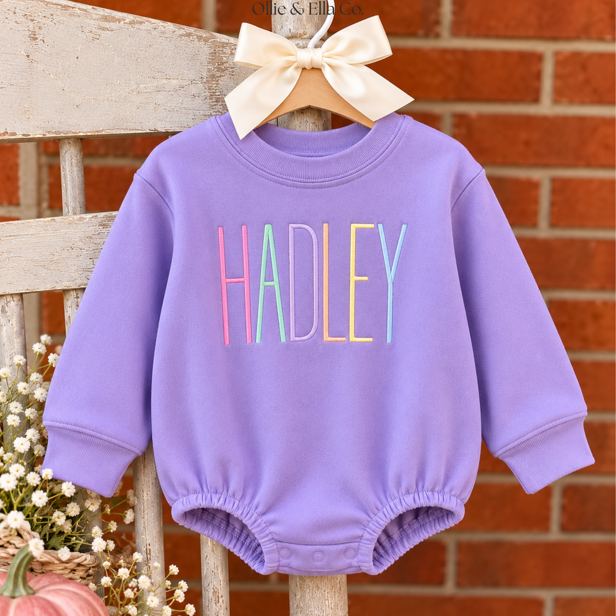 A lavender baby long-sleeve romper with the name 'HADLEY' printed in colorful pastel letters, hanging on a wooden hanger with a cream bow against a brick wall and rustic wooden chair backdrop.