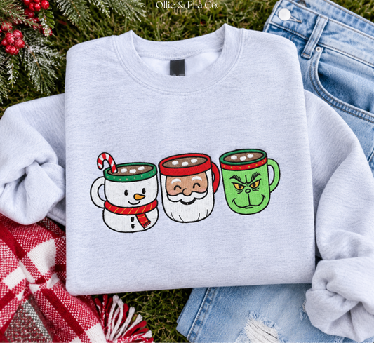 A light gray sweatshirt laid flat on a grassy surface with holiday styling props, including pine branches, red berries, a red-and-white plaid blanket, and blue jeans. The sweatshirt has a printed graphic of three mug faces: a snowman mug with a candy cane, a Santa mug, and a green Grinch-style mug.