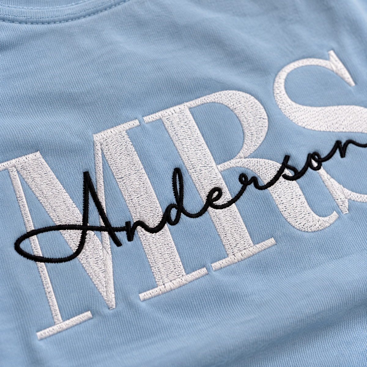 Close-up of a light blue embroidered T-shirt with large white block letters spelling 'MRS' and a black cursive script name 'Anderson' across the front.
