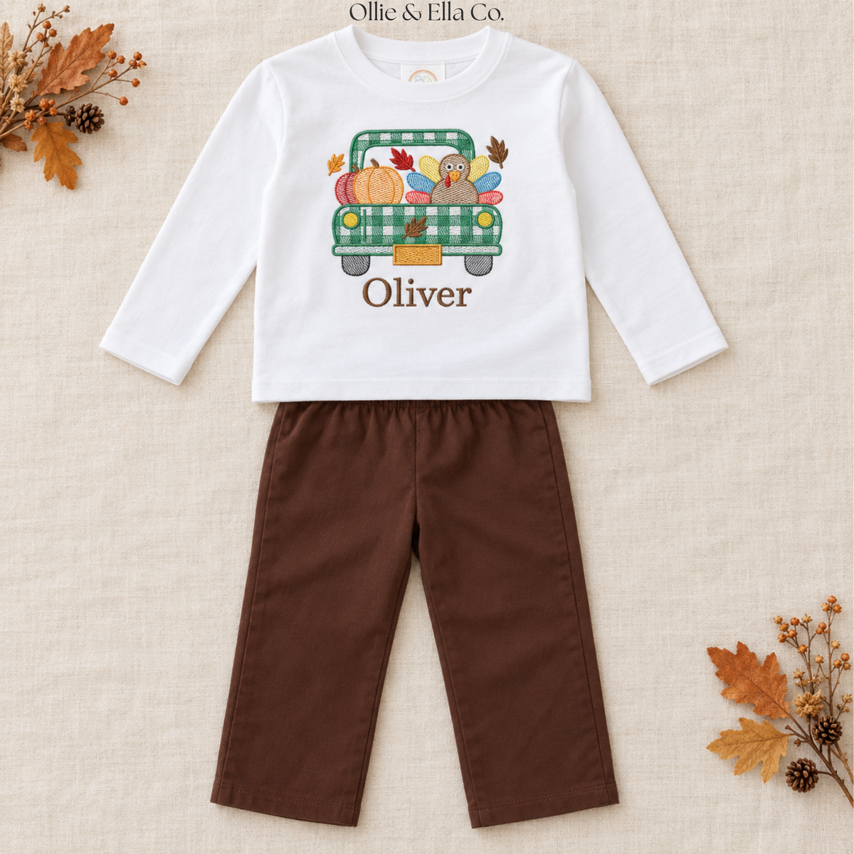 A toddler boy's two-piece outfit set by Ollie & Ella Co. featuring a white long-sleeve shirt with a green plaid truck graphic and the name 'Oliver' printed on the front, paired with brown pants, laid flat on a textured beige fabric background with autumn leaf props.