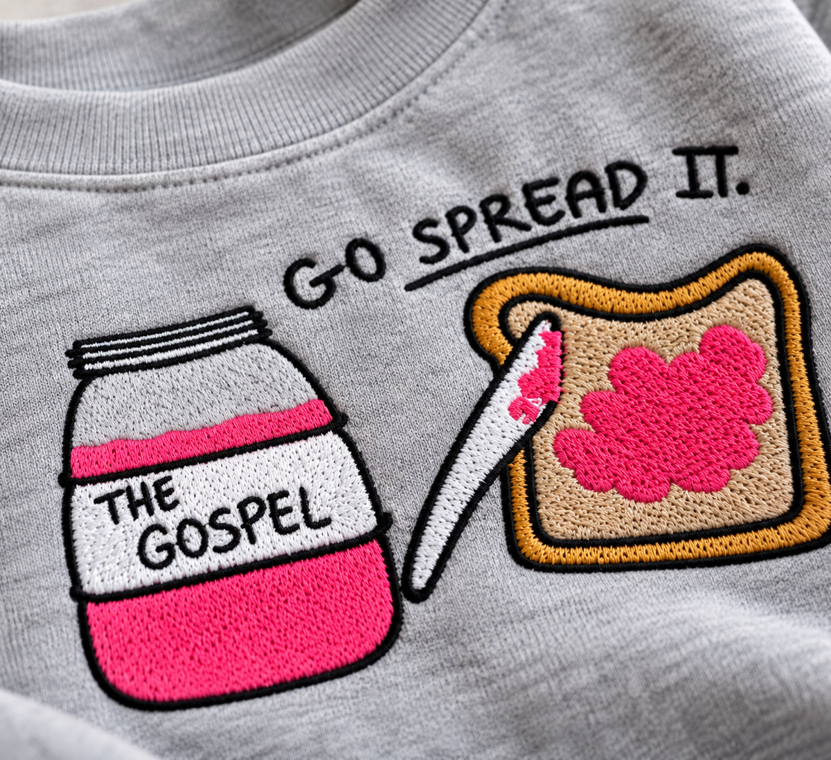 Close-up of a light gray crewneck sweatshirt featuring a colorful embroidered graphic of a jar labeled 'THE GOSPEL' with pink jam, a knife, and a slice of toast with pink jam, plus the text 'GO SPREAD IT.'.