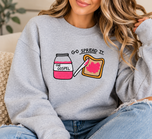 A woman wearing a light heather gray crewneck sweatshirt with a graphic of a pink jam jar labeled 'THE GOSPEL' and a slice of toast with pink spread, captioned 'GO SPREAD IT.' She is seated indoors on a textured couch.