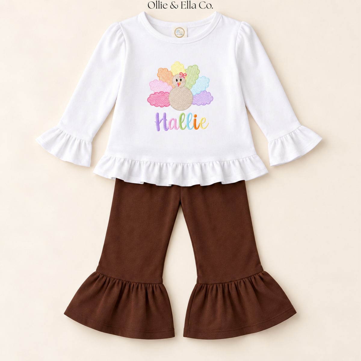 A toddler girl's two-piece outfit set featuring a white long-sleeve top with ruffled cuffs and hem, decorated with a colorful turkey graphic and the word 'Hattie' in pastel lettering. It is paired with brown flared pants that have matching ruffled hems, displayed on a light beige background.