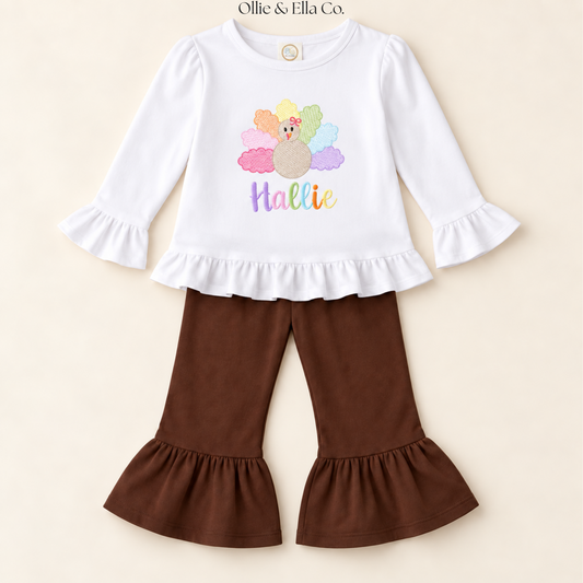 A toddler girl's two-piece outfit set featuring a white long-sleeve top with ruffled cuffs and hem, decorated with a colorful turkey graphic and the word 'Hattie' in pastel lettering. It is paired with brown flared pants that have matching ruffled hems, displayed on a light beige background.