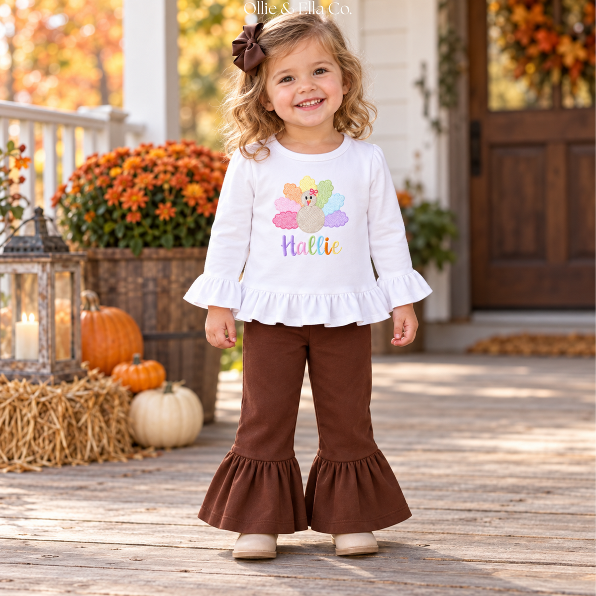A young girl stands on a wooden porch wearing a white long-sleeve ruffled top with a colorful turkey graphic and the word 'Halloween' on the front, paired with brown flared pants. Pumpkins, flowers, and autumn decorations are visible in the background.
