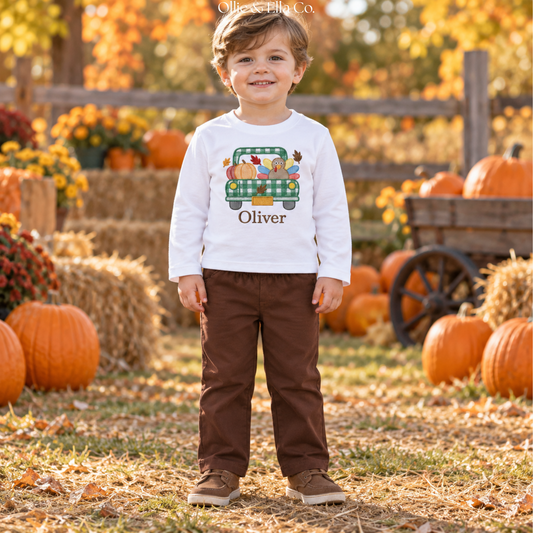 A young boy stands in a pumpkin patch wearing a white long-sleeve shirt with a green plaid truck graphic and the name 'Oliver' printed on the front, paired with brown pants and brown shoes.