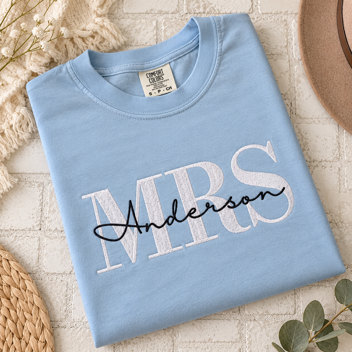 A light blue folded t-shirt with 'MRS Anderson' printed on the front in white and black, styled on a white brick background with decorative props.